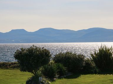 A garden with bushes and grass overlooking a body of water and mountains at Seafront Holiday Home 2 in Pwllheli