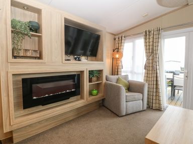 A living room with an armchair a wall-mounted television an electric fireplace and sliding glass doors with curtains at Seafront Holiday Home 2 in Pwllheli
