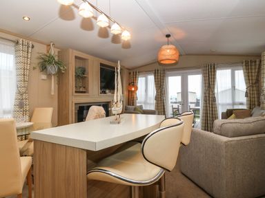 An open living room and kitchen area with bar stools a sofa a glass dining table and large windows with curtains at Seafront Holiday Home 2 in Pwllheli