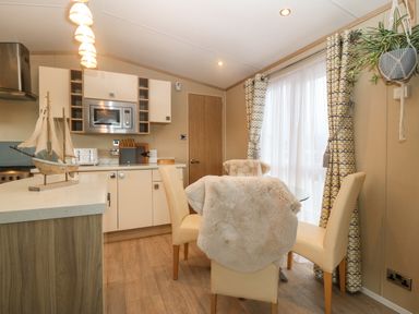 A dining area with four chairs around a glass table next to a kitchen with cabinets and a microwave at Seafront Holiday Home 2 in Pwllheli