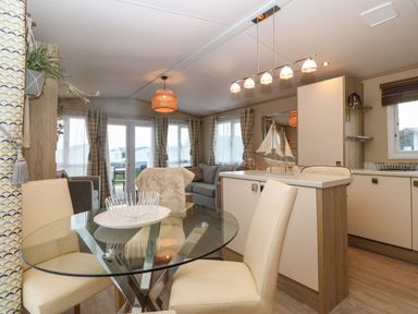 A dining area with a glass table and cream chairs next to a kitchen island and a seating area with grey sofas at Seafront Holiday Home 2 in Pwllheli