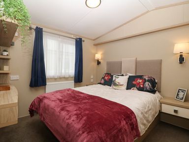 A bedroom with a double bed red blanket floral pillows two wall lamps a window with blue curtains and shelves on the wall at Seafront Holiday Home 2 Pwllheli