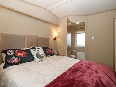 A bedroom with a bed linens with floral pillows a wall light and an open door to a bathroom at Seafront Holiday Home 2 in Pwllheli