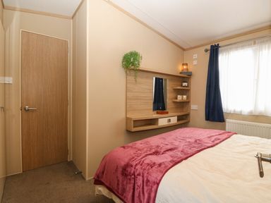 A bedroom with a wooden door a bed with a pink blanket a wall-mounted shelf with a mirror and plants a window with blue curtains at Seafront Holiday Home 2 in Pwllheli