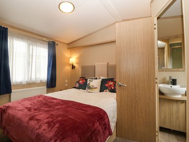 A bedroom with a bed covered with red and cream bedding blue curtains on a window and a door leading to a bathroom at Seafront Holiday Home 2 in Pwllheli