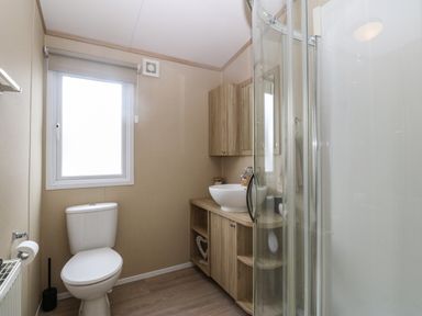 A bathroom with a toilet a window a wooden vanity unit with a sink and a curved glass shower enclosure at Seafront Holiday Home 2 Pwllheli