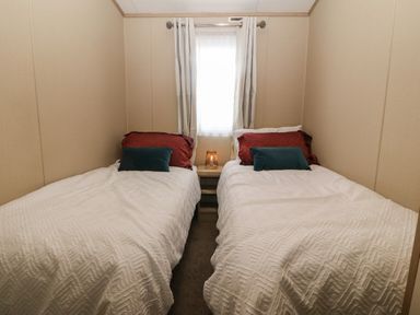 A bedroom with two single beds separated by a nightstand with a lamp and a window with curtains at Seafront Holiday Home 2 in Pwllheli