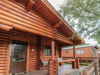 The exterior porch of a wooden log cabin with a door windows chairs and a railing at Welcome Hjem Log Cabin in Morpeth near Felton