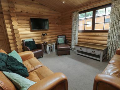 A living room with leather sofas and armchairs a coffee table under a window and a wall-mounted television at Welcome Hjem Log Cabin Morpeth near Felton