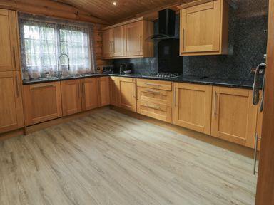 A kitchen with wooden cabinets black countertops a gas stove and a window with lace curtains at Welcome Hjem Log Cabin Morpeth near Felton
