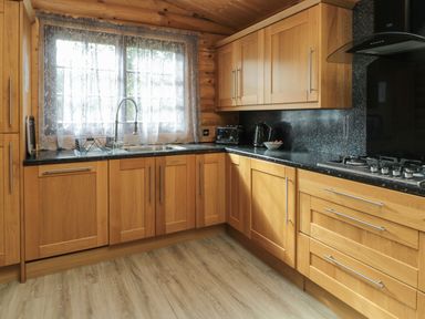 A kitchen with wooden cabinets black countertops a window with lace curtains and a gas stove at Welcome Hjem Log Cabin Morpeth near Felton