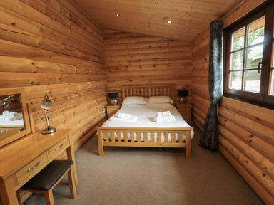 A bedroom with a wooden bed, two bedside tables with lamps, a wooden desk with a lamp, and a window with curtains at Welcome Hjem Log Cabin in Morpeth near Felton