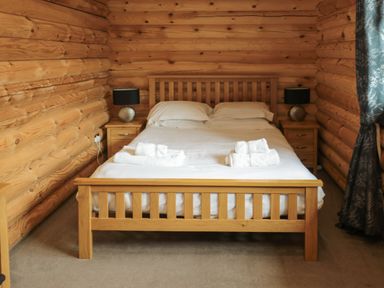 A bedroom with a wooden bed frame white bedding bedside tables with lamps and wooden log walls at Welcome Hjem Log Cabin Morpeth near Felton