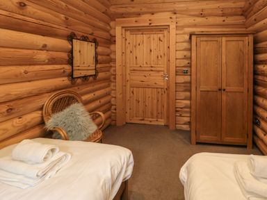A bedroom with two beds with white linens a wooden wardrobe a wooden door a chair with a cushion and a wall mirror at Welcome Hjem Log Cabin Morpeth near Felton