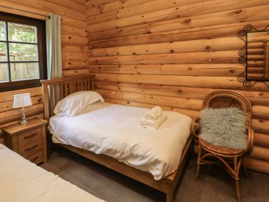 A bedroom with two beds a nightstand with a lamp a window and a chair with a cushion at Welcome Hjem Log Cabin Morpeth near Felton