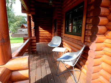 A wooden log cabin porch with two woven chairs and a small table at Welcome Hjem Log Cabin in Morpeth near Felton
