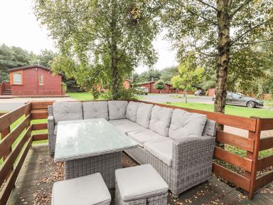 An outdoor seating area with a wicker sofa and glass table on a wooden deck surrounded by trees and cabins at Welcome Hjem Log Cabin Morpeth near Felton