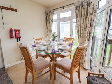 A dining area with a glass table and four wooden chairs next to large windows with curtains at Eden Lodge in Castlewigg near Whithorn