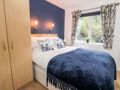 A bedroom with a double bed blue blanket and pillows a wooden wardrobe a window with curtains and a nightstand lamp at Eden Lodge in Castlewigg near Whithorn