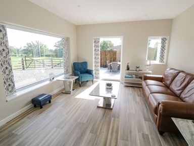 A living room with a brown leather sofa a blue armchair a coffee table a side table with a lamp and books large windows with patterned curtains and a door leading to an outdoor patio at Southend in Welland