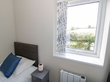 A bedroom corner with a bed with blue pillow a bedside table with a small lamp a window with patterned curtains and a radiator below the window at Southend in Welland