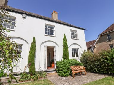 An exterior view of a white two-story house with tall windows and an open door with a wooden bench on a stone patio at Charmouth Lodge in Charmouth