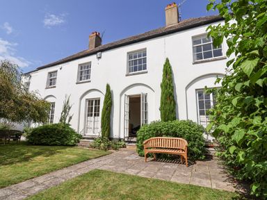A white two-story house with multiple windows and doors opening to a garden with shrubs and a wooden bench at Charmouth Lodge in Charmouth