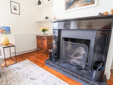 A corner of a room with a black fireplace a wooden cabinet with flowers on it a small table with a yellow lamp and a light colored rug on a wooden floor at Charmouth Lodge in Charmouth