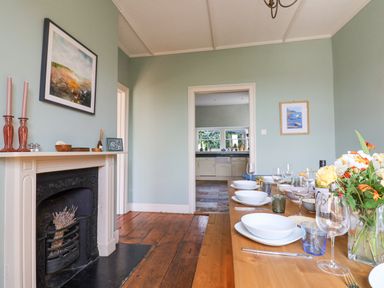 A dining room with a wooden floor fireplace and a set table next to an open doorway to a kitchen at Charmouth Lodge in Charmouth