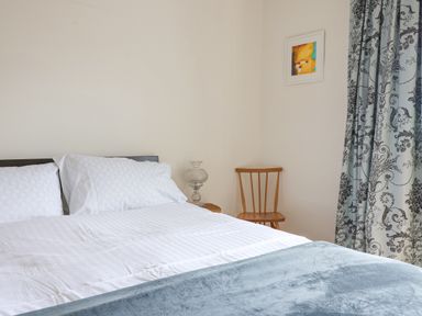 A bedroom with a bed with white linens a wooden chair a bedside table with a lamp patterned curtains and a framed painting on the wall at Charmouth Lodge in Charmouth