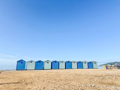 A row of blue and light blue beach huts on a sandy beach with a clear sky