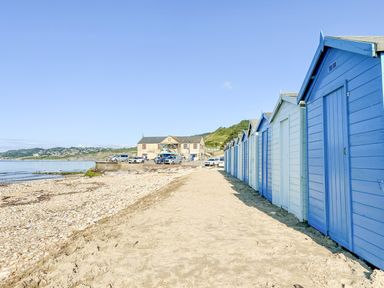 A row of blue and white beach huts along a sandy beach with a building and cars in the background at Charmouth Lodge in Charmouth