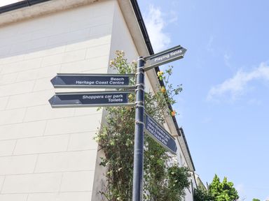 A signpost with directions to beach heritage coast centre shoppers car park toilets playing fields tourist information point library internet cafe parish council office next to a white building with plants at Charmouth Lodge in Charmouth
