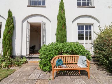 A wooden bench with a blanket and books on a stone patio in front of a white building with open doors and green bushes at Charmouth Lodge in Charmouth
