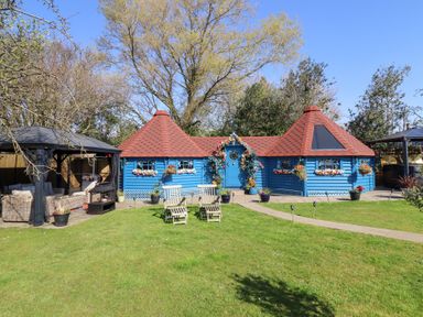 A blue wooden cabin with red roofs surrounded by flower pots and two wooden chairs on a lawn at Applejacks Orchard in Holywell