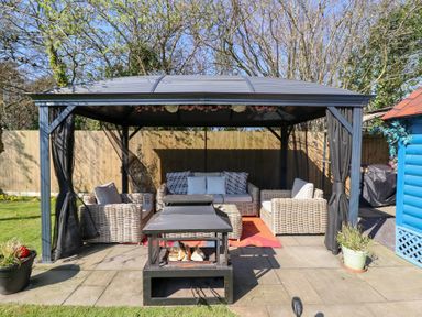 An outdoor seating area with wicker chairs and a sofa under a metal gazebo with a fire pit in front at Applejacks Orchard in Holywell