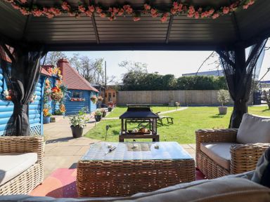 A covered outdoor seating area with wicker furniture looking out to a garden with a fire pit and a blue building at Applejacks Orchard in Holywell