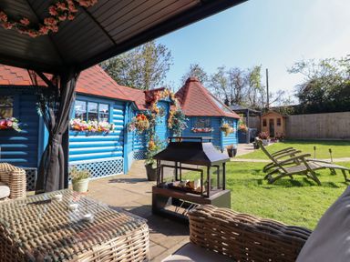 An outdoor seating area with wicker furniture and a fire pit near blue wooden cabins with flower decorations at Applejacks Orchard in Holywell