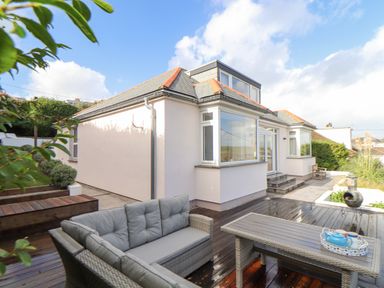 An outdoor patio with wicker sofa and table with tea set next to a house with large windows at Higher Bolenna in Perranporth