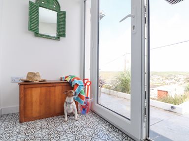 A small dog sitting on patterned tile floor next to a wooden bench with a hat and colorful towel beside a glass door with outdoor view at Higher Bolenna in Perranporth