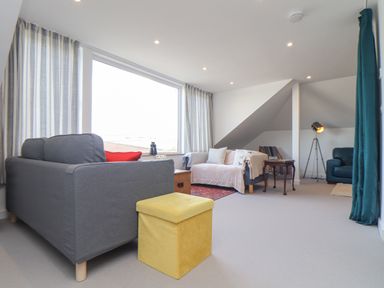 A living room with sofas a yellow ottoman a floor lamp and a large window at Higher Bolenna in Perranporth