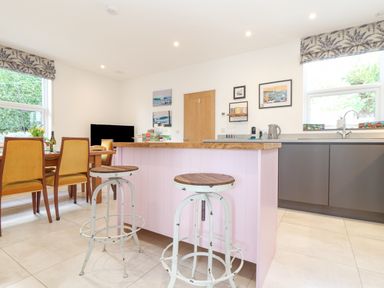 A kitchen with a wooden island and two stools a dining table with chairs a sink under a window and pictures on the wall at Higher Bolenna in Perranporth