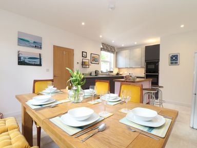 A dining table set with plates bowls wine glasses and cutlery next to a kitchen with stools at Higher Bolenna in Perranporth