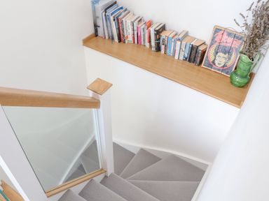 A staircase with grey carpet and wooden handrail and a shelf with books a green jug with dried flowers and a framed picture at Higher Bolenna in Perranporth