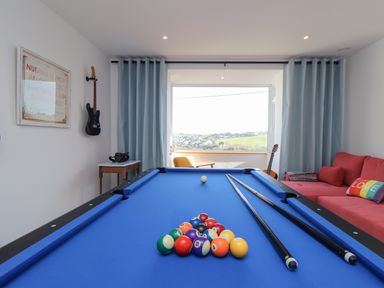 A game room with a blue pool table two pool cues and a rack of balls a red couch with pillows a guitar on a stand and a large window at Higher Bolenna in Perranporth