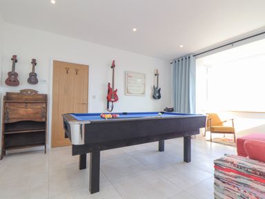 A game room with a pool table guitars on the wall a wooden cabinet a chair and a window with curtains at Higher Bolenna in Perranporth