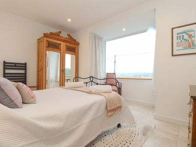 A bedroom with a bed, pillows, a wooden wardrobe with mirrors, a chair near a large window, a framed picture on the wall, and a rug on the floor at Higher Bolenna in Perranporth