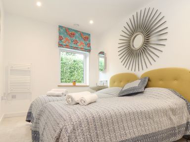 A bedroom with two single beds with patterned bedcovers and pillows a window with a floral roman blind a washbasin and a decorative sunburst mirror on the wall at Higher Bolenna in Perranporth