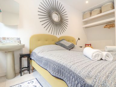 A bedroom with a double bed with a patterned bedspread two rolled towels a decorative mirror shelves with storage bins and a small sink at Higher Bolenna in Perranporth