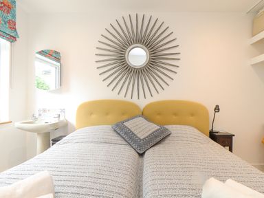 A bedroom with twin beds yellow headboards a round decorative mirror a bedside table and a sink at Higher Bolenna in Perranporth